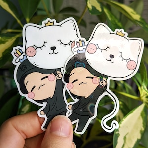 May include: Two black and white cartoon stickers of people holding white cat balloons with crowns. The balloons have pink cheeks and closed eyes. The people are wearing black shirts and have pink cheeks. The stickers are on a hand.