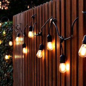 Puede incluir: Una guirnalda de luces para exteriores con bombillas estilo Edison, casquillos y cables negros, que emiten un cálido resplandor amarillo. Las luces están suspendidas a lo largo de una valla de listones de madera verticales.