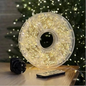 Może przedstawiać: Przezroczysta plastikowa szpula owinięta cienkim miedzianym drutem z licznymi małymi diodami LED o ciepłej białej barwie. Czarny zasilacz z dwubolcową wtyczką oraz mały prostokątny pilot z przyciskami znajdują się przed szpulą.