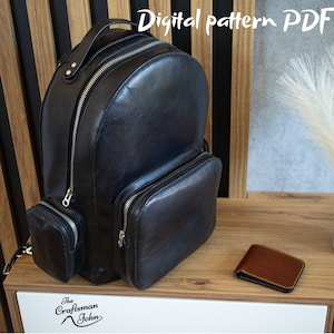 Può includere: Uno zaino in pelle nera con più tasche e cerniere. Lo zaino è appoggiato su una superficie in legno con un piccolo portafoglio in pelle marrone e un vaso con erba della pampa bianca.