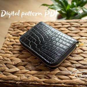 Può includere: Un portafoglio nero strutturato con cerniera dorata, appoggiato su una superficie di vimini intrecciato. Il portafoglio presenta un motivo a rilievo simile alla pelle di rettile. Il testo "Digital pattern PDF" è visibile. Il logo The Craftsman John è in basso a destra.
