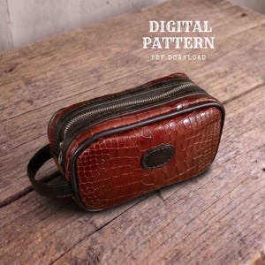 Può includere: Una borsa da toilette rettangolare marrone con texture effetto coccodrillo, chiusura a zip marrone scuro e manico laterale. Il testo DIGITAL PATTERN PDF DOWNLOAD è visibile sopra la borsa. Presenta una toppa ovale marrone scuro sul davanti.