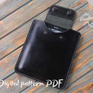 Puede incluir: Una funda de cuero negro para Kindle con cierre de solapa y borde cosido. La funda está abierta y el Kindle es visible en el interior. La funda está sobre una superficie de madera con una mancha azul y marrón.