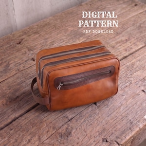 Pode incluir: Uma nécessaire de couro bege com dois compartimentos principais com zíper e um bolso frontal menor com zíper e acabamento em marrom escuro. Uma alça lateral está fixada na lateral. O texto DIGITAL PATTERN PDF DOWNLOAD está visível.