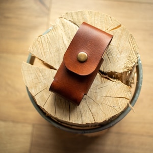 Może przedstawiać: Brązowe skórzane etui z klapką i mosiężnym zapięciem na guzik. Etui jest prostokątne i wygląda na ręcznie robione. Spoczywa na jasnym przekroju pnia drzewa, z drewnianą podłogą w tle.