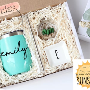 May include: A gift box filled with a teal tumbler with the name "emily" on it, a small glass terrarium with a succulent plant, a white dish with the letter "E" on it, and a clear glass straw. The box is filled with white shredded paper. The box is labeled with the text "more colors available". A small white box with a green plant is visible in the background. A yellow sun with a smiling face and the text "you are my sunshine" is visible in the bottom right corner.