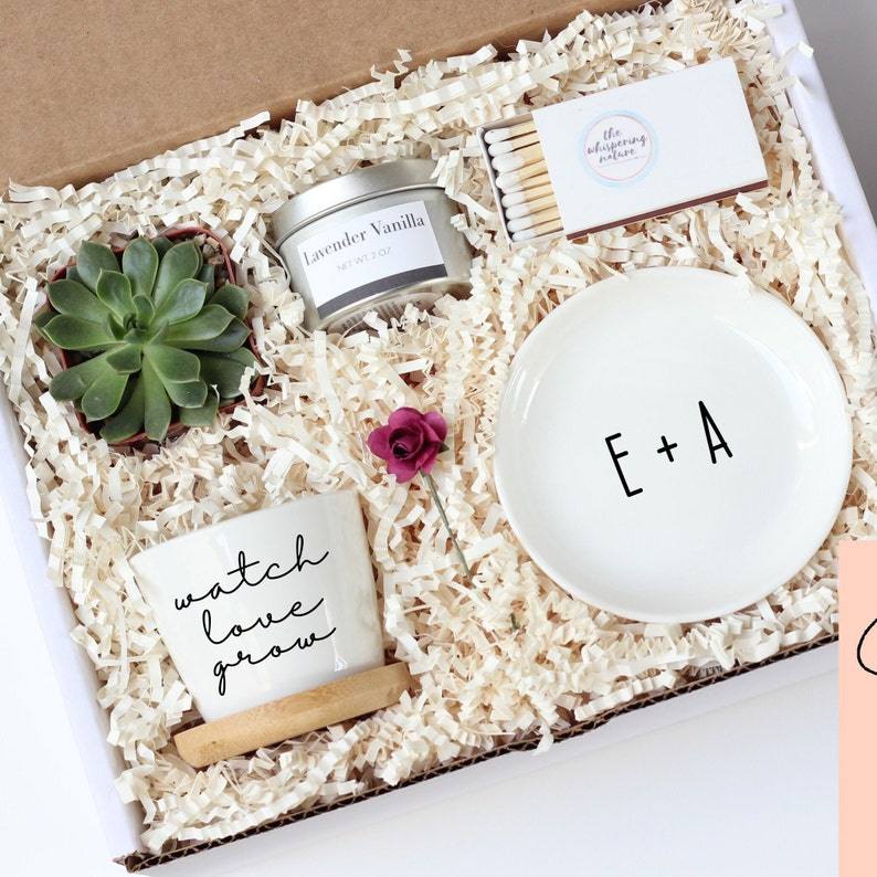 Puede incluir: Una caja de regalo llena de una planta suculenta, una vela peque&ntilde;a, un plato con las iniciales E+A, una maceta peque&ntilde;a con las palabras "watch love grow" y una caja de cerillas. La caja est&aacute; llena de papel blanco triturado.