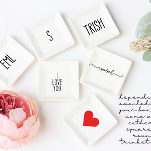 May include: Six white square trinket dishes with various designs. The designs include the words "EML", "S", "TRISH", "I LOVE YOU", "isabel", and a red heart. The dishes are arranged on a white surface.