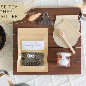 Op de afbeelding: Een houten dienblad met een bruine papieren zak met losse thee, een klein potje honing, een theefilter van mousseline en een houten honingroerder. De tekst "Loose Tea" staat op de zak gedrukt.