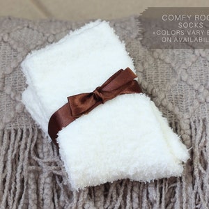 Op de afbeelding: Een paar witte, pluche sokken gebonden met een bruin lint. De sokken liggen op een bruin gebreid deken met franjes. Comfortabele kamer sokken *Kleuren vari&euml;ren afhankelijk van de beschikbaarheid.
