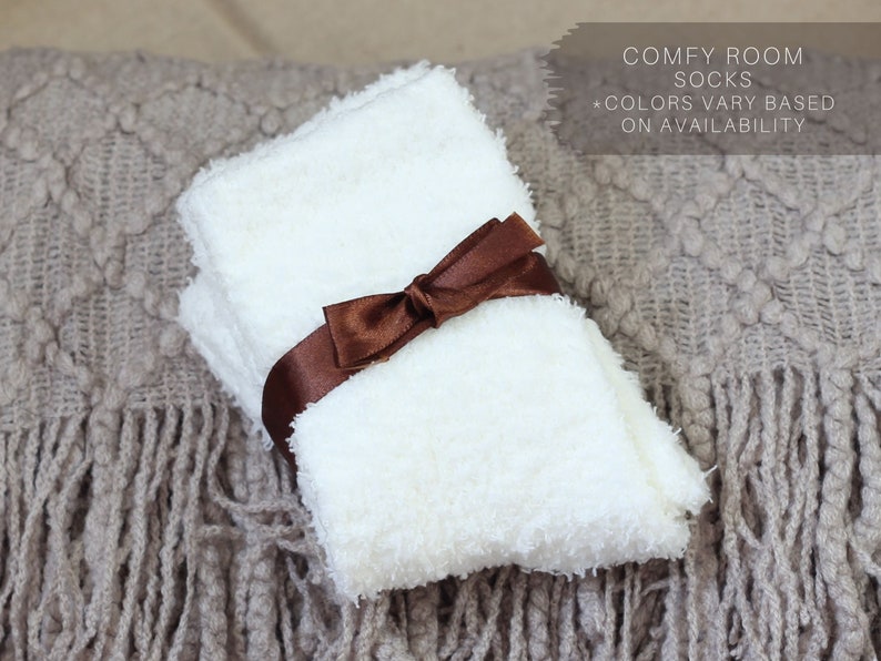 Op de afbeelding: Een paar witte, pluche sokken gebonden met een bruin lint. De sokken liggen op een bruin gebreid deken met franjes. Comfortabele kamer sokken *Kleuren vari&euml;ren afhankelijk van de beschikbaarheid.