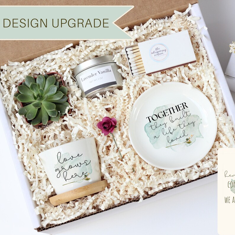 Puede incluir: Una caja de regalo llena de art&iacute;culos para un regalo de inauguraci&oacute;n de la casa. La caja contiene una peque&ntilde;a planta suculenta, una vela, una peque&ntilde;a maceta de cer&aacute;mica con el texto "el amor crece aqu&iacute;", un peque&ntilde;o plato de cer&aacute;mica con el texto "Juntos construyeron una vida que amaron", y una caja de cerillas.