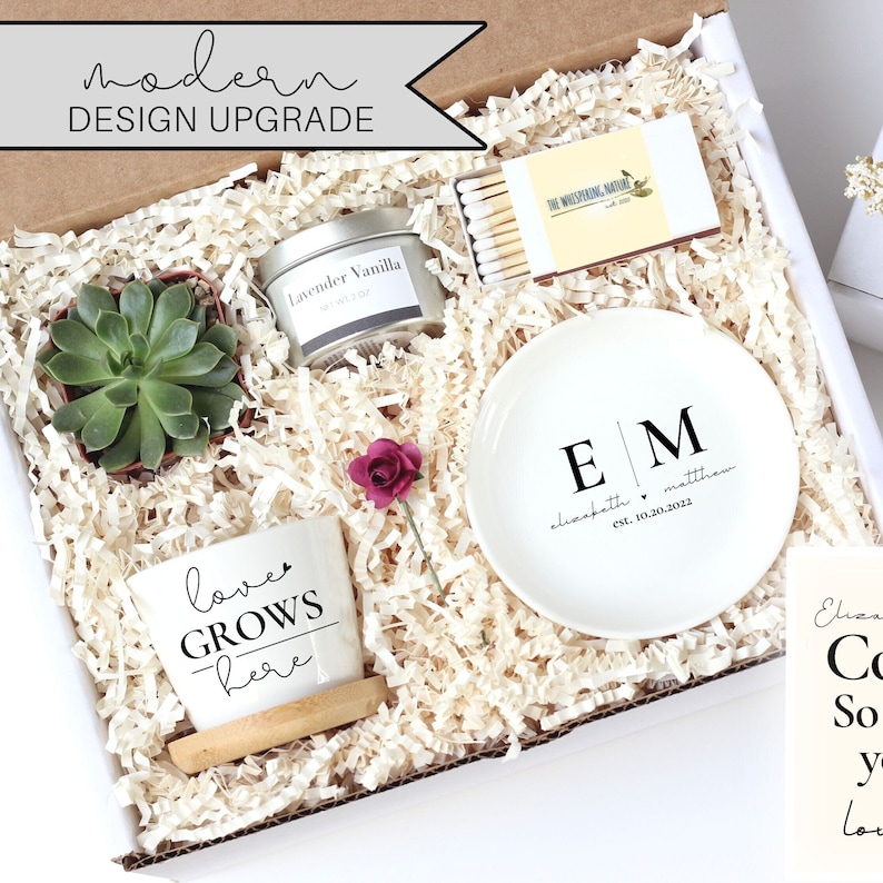 May include: A white gift box filled with a variety of items, including a small succulent plant, a candle, a small dish with the initials "EM" and the text "elizabeth matthew est. 10.20.2022", a small white ceramic mug with the text "love grows here", and a box of matches. The box is filled with white shredded paper.
