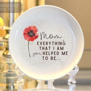 Op de afbeelding: Witte keramische sieradenschotel met een rode klaproos en de tekst "Mam, alles wat ik ben, heb je me geholpen te zijn..." De schotel heeft een diameter van 11,4 cm en staat op een heldere standaard.