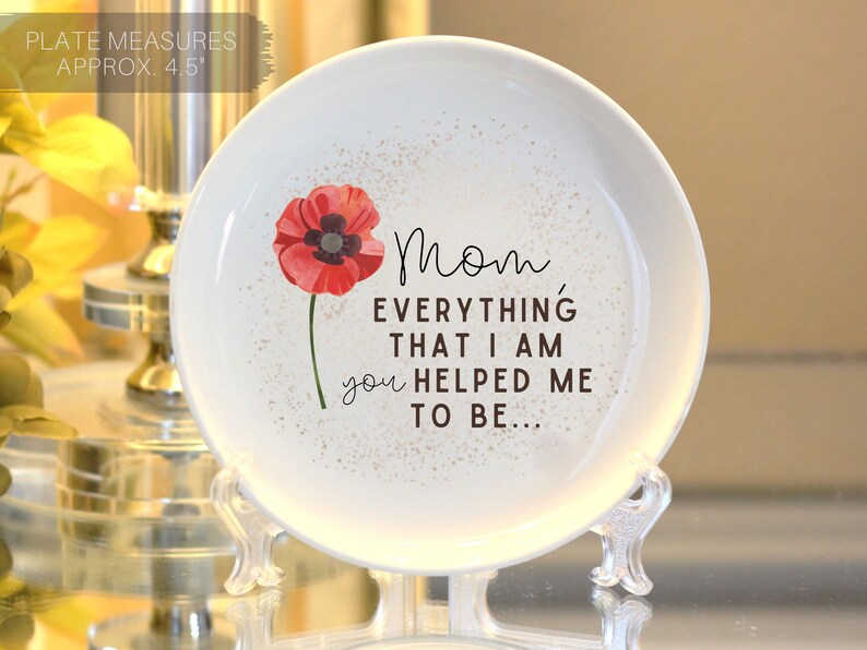 Op de afbeelding: Witte keramische sieradenschotel met een rode klaproos en de tekst "Mam, alles wat ik ben, heb je me geholpen te zijn..." De schotel heeft een diameter van 11,4 cm en staat op een heldere standaard.