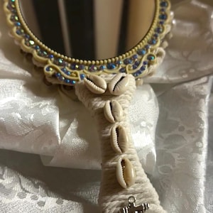 May include: A decorative hand mirror with a round, cream-colored frame adorned with iridescent blue and clear crystals. The handle is wrapped in cream-colored rope and decorated with seashells and a silver anchor charm.