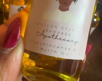May include: A glass bottle with a yellow liquid inside. The label reads "African Goddess Apothecary Frankincense & Myrrh Oil". The label also features a drawing of a woman with butterflies in her hair.