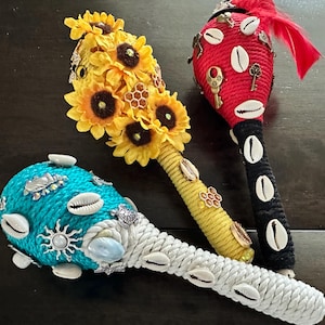 Op de afbeelding: Drie maracas met verschillende ontwerpen. Een maraca is bedekt met gele zonnebloemen en honingraat, een andere is rood met witte schelpen en sleutels, en de laatste is turquoise met witte schelpen en een zon.