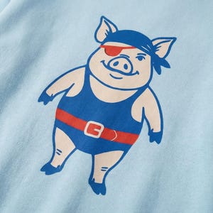 Op de afbeelding: Lichtblauw T-shirt met een cartoon varkensillustratie. Het varken draagt een blauw badpak, rode riem en ooglapje, met een blauwe bandana. Het varken heeft een lachend gezicht. Het ontwerp is gedrukt op een lichtblauwe achtergrond.