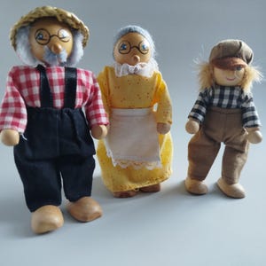 Könnte beinhalten: Drei Holzpuppen, die eine Familie darstellen. Der Vater trägt einen Strohhut, ein rot-weiß kariertes Hemd, eine schwarze Latzhose und Holzschuhe. Die Mutter trägt ein gelbes Kleid und eine weiße Schürze. Das Kind trägt eine Mütze, ein kariertes Hemd und eine braune Hose.