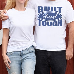 May include: Two people wearing white t-shirts. One shirt has the text "BUILT Dad TOUGH" in blue. The other person is wearing a plain white t-shirt and light blue jeans. The background is a red wooden wall.