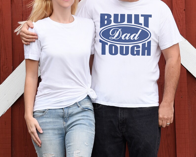 May include: Two people wearing white t-shirts. One shirt has the text "BUILT Dad TOUGH" in blue. The other person is wearing a plain white t-shirt and light blue jeans. The background is a red wooden wall.