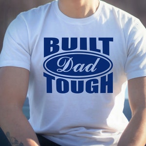 May include: White t-shirt with the text "BUILT Dad TOUGH" in blue. The text is arranged in a stacked format, with "Dad" inside an oval design. The shirt is short-sleeved and appears to be made of cotton.