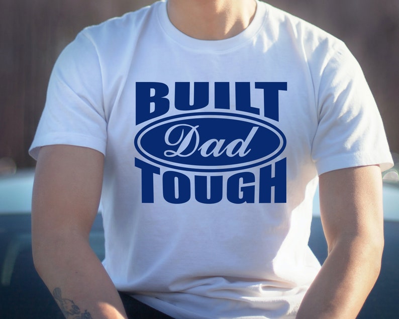 May include: White t-shirt with the text "BUILT Dad TOUGH" in blue. The text is arranged in a stacked format, with "Dad" inside an oval design. The shirt is short-sleeved and appears to be made of cotton.