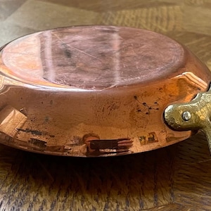 May include: A small, copper saucepan with a rounded base and a brass handle. The pan has a shiny, reflective surface and is designed for cooking. The handle is attached with rivets. This vintage cookware is ideal for a kitchen.