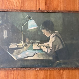 May include: Framed vintage print depicting a young person seated at a desk, illuminated by a desk lamp. The print features a boy writing, with a soft color palette of blues, greens, and browns. The frame is a warm, light brown color.