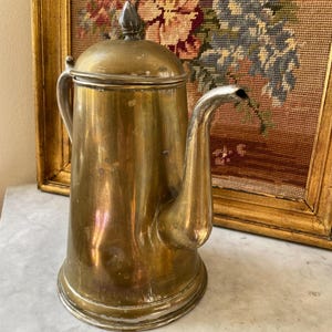 Op de afbeelding: Een vintage messing koffiepot met een gebogen tuit en een deksel met een puntige knop. De pot heeft een handvat en staat op een marmeren oppervlak. De achtergrond omvat een ingelijste wandtapijt met bloemmotieven.