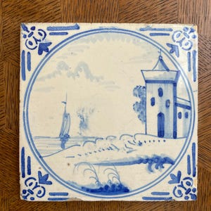 May include: Square ceramic tile with a hand-painted seascape scene in shades of blue. The design features a sailboat, a building, and a decorative border. The tile is likely vintage and shows signs of wear.