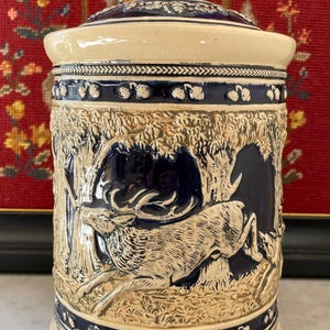 Può includere: Un vaso in ceramica beige e blu navy con coperchio. Il vaso presenta un disegno a rilievo di un cervo in una scena di foresta. Il coperchio è sormontato da un pomello blu scuro con pois bianchi. È un oggetto decorativo.