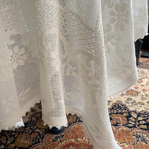 Peut inclure: Une nappe blanche en tissu ajouré avec des motifs floraux et géométriques complexes. Les bords sont finis avec une bordure festonnée. Le tissu recouvre une table, mettant en valeur le savoir-faire et la texture du tissu.