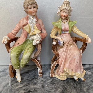 Puede incluir: Un par de figuras antiguas de porcelana que representan a un hombre y una mujer sentados en sillas. El hombre viste un abrigo rosa y pantalones verdes, mientras que la mujer lleva un vestido rosa y una chaqueta verde. Ambas figuras tienen rasgos faciales detallados.