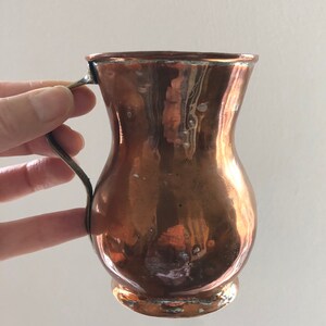 May include: A copper jug with a flared spout and a handle. The jug has a worn, aged look.