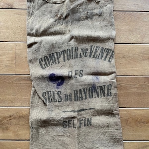 May include: A vintage burlap sack with the text "COMPTOIR DE VENTE DES SELS DE BAYONNE SEL FIN" printed in black on the front.