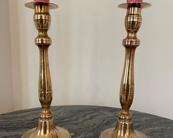 May include: Two antique brass candlesticks with pink candles. The candlesticks have a classic design with a round base, a slender stem, and a small dish to hold the candle. The candlesticks are placed on a marble surface.