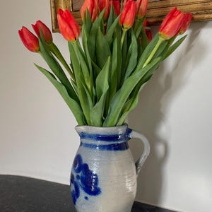 May include: A display of red tulips with green foliage in a blue and white ceramic jug. The jug features a handle and blue floral patterns. The flowers are arranged in front of a framed picture on the wall.