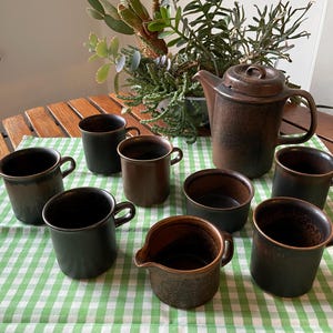 Op de afbeelding: Een set van bruine keramische mokken en een bijpassende koffiepot op een bruin-groen geruit tafelkleed. De mokken en koffiepot hebben een getextureerde, rustieke uitstraling.