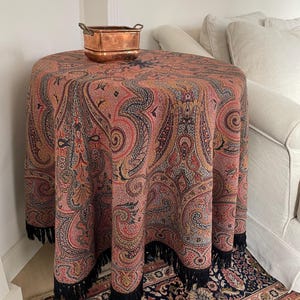 May include: A round table covered with a paisley patterned tablecloth in shades of red, orange, and blue. The tablecloth has a black fringe. A copper container with handles sits on top of the table.
