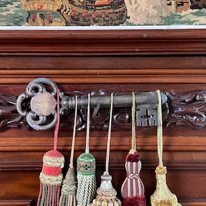 May include: A large, weathered metal skeleton key mounted horizontally on a dark wood surface, serving as a decorative rack. Five distinct fabric tassels in shades of red, green, gold, and beige hang from the key by thin, braided cords.