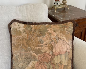 May include: A decorative tapestry pillow featuring a pastoral scene with figures in period clothing. The pillow has a brown fringe border and is set against a neutral-toned sofa. The artwork depicts a man and two women in a garden setting.