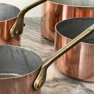 Op de afbeelding: Close-up van koperen pannen met messing handvatten. De pannen hebben een glanzende, roodbruine buitenkant en een zilveren binnenkant. De handvatten zijn gebogen en met klinknagels bevestigd. De pannen zijn van verschillende maten en vormen.