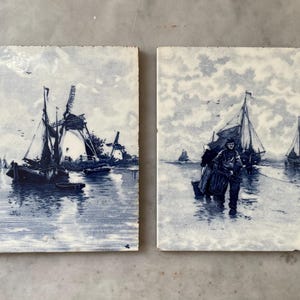 May include: Two square ceramic tiles with blue and white nautical scenes. One tile depicts boats and windmills, the other shows figures wading in water near sailboats. The tiles have a vintage, hand-painted aesthetic.