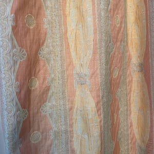 May include: Close-up of a peach-colored fabric with vertical stripes and ornate detailing. The design features alternating patterns of light peach, cream, and gold, creating a textured and elegant appearance. The fabric has a subtle sheen.