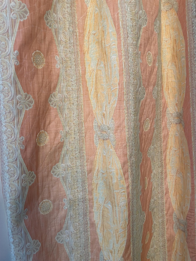 May include: Close-up of a peach-colored fabric with vertical stripes and ornate detailing. The design features alternating patterns of light peach, cream, and gold, creating a textured and elegant appearance. The fabric has a subtle sheen.