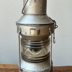 Op de afbeelding: Een zilverkleurige metalen lantaarn met een cilindrisch lichaam en een middengedeelte van helder glas. De lantaarn heeft een getrapte bovenkant met een handvat, een brede basis en verticale metalen steunen die de boven- en onderkant verbinden.