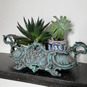 May include: An ornate, antique-style planter with a verdigris finish, holding two succulents. The planter has intricate scrollwork and handles. One succulent is in a small blue and white pot.