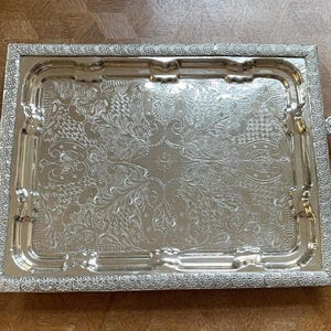 May include: A rectangular silver-toned serving tray featuring an ornate embossed floral and scrollwork pattern across the center surface. The tray has a wide, textured border with a repeating flower motif and two decorative curved metal handles.